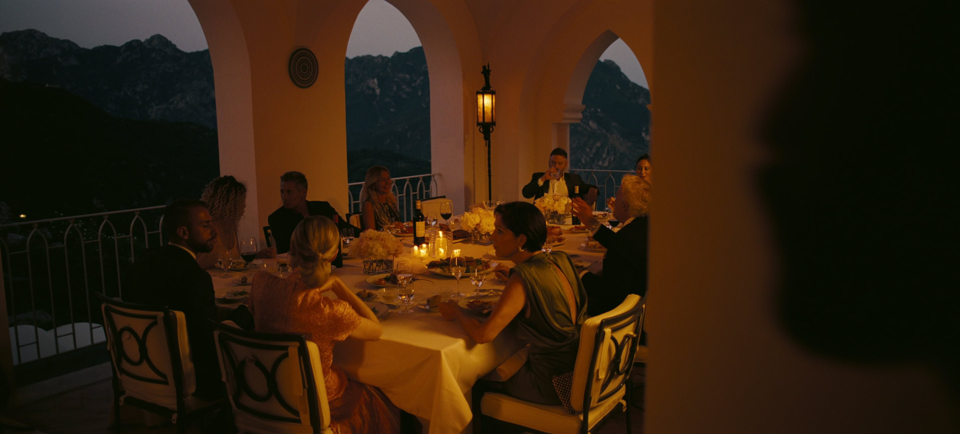 a group of people sit around a dinner table on a terrace overlooking a mountain vista. the table is set with white linens, candles, and glasses of wine. they are all dressed in formal attire. the setting is elegant and romantic.  the terrace is bathed in the warm glow of sunset.  the people are smiling and laughing, enjoying each other's company.  the view of the mountains is breathtaking.