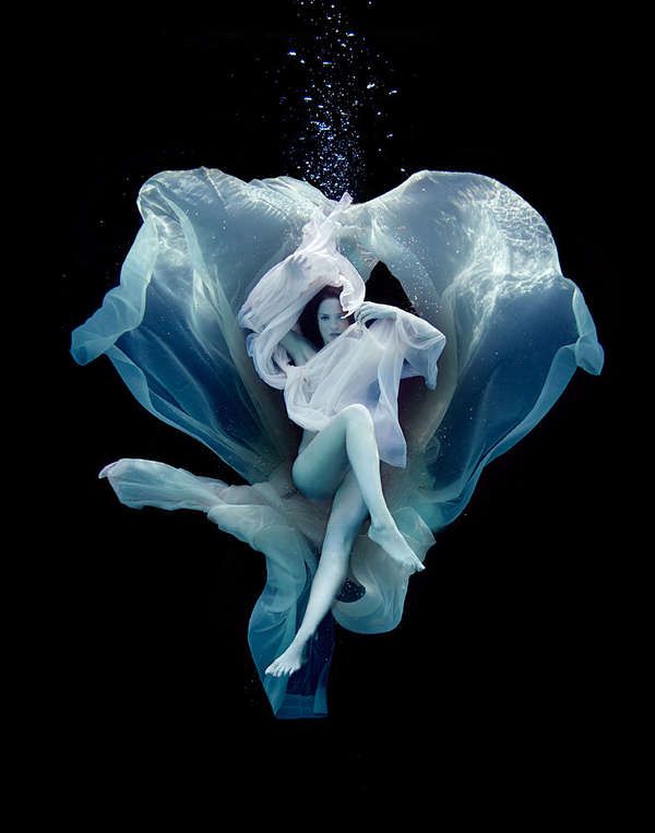 a woman in a flowing white dress is submerged underwater. she is surrounded by swirling blue fabric, giving the impression of a watery vortex. the image is dark and mysterious, creating a sense of elegance and ethereal beauty. the flowing fabric and underwater setting contribute to a sense of fantasy and artistic expression.