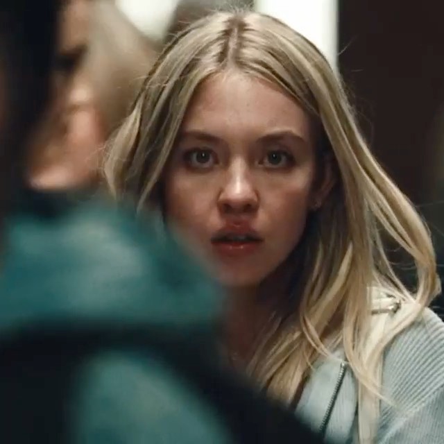 the image is a closeup shot of a young blonde woman with a look of fear and anxiety on her face.  her expression suggests she is frightened or worried about something. her blonde hair is long and slightly tousled. she's wearing a light blue sweater and appears to have a shoulder bag. the background is blurry but appears to be an indoor location, possibly a school hallway. the focus is primarily on the woman's face and expression, conveying a sense of suspense and tension, suggesting a scene from a thriller or drama film. a blurred figure in teal is partially visible in the foreground, adding to the sense of mystery.