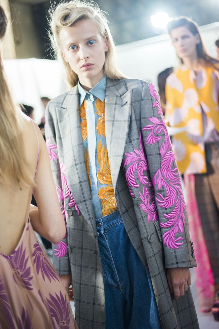 closeup shot of a fashion model backstage at a runway show. the model is wearing a grey plaid blazer with pink floral embroidery on the sleeves, a light blue shirt with an orange floral pattern, and blue jeans. her hair is styled in a retro updo. another model in a yellow dress is visible in the background.  the overall aesthetic is high fashion and polished.