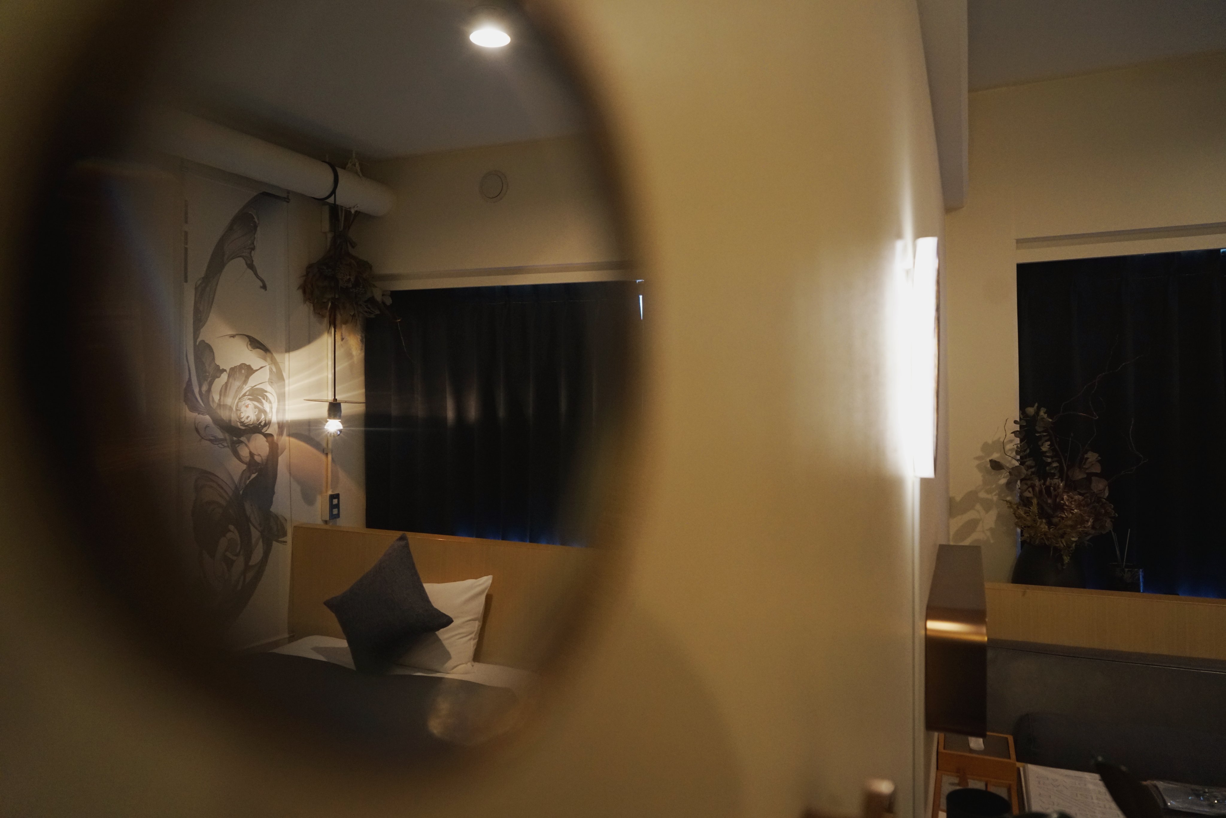 a bedroom is shown through a peephole, creating a voyeuristic feel. the room is decorated in a minimalist style with a neutral color palette. the bed is made, and a single pillow rests on top.  the warm lighting from the lamp creates a cozy atmosphere.