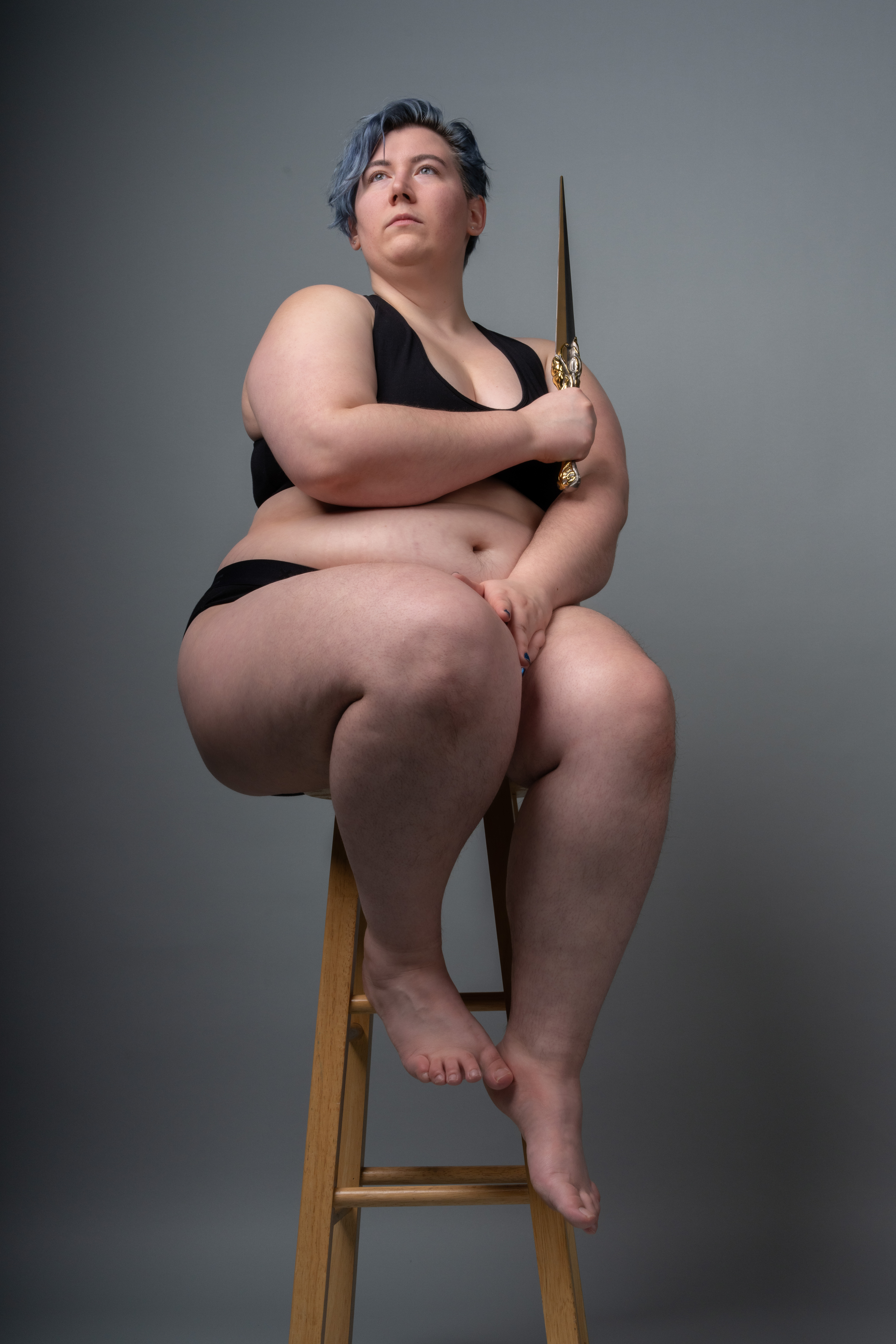 a plussize woman with blue hair is sitting on a wooden stool in a studio setting. she is wearing black lingerie and holding a sword. she is looking up and appears contemplative. the background is a solid grey color.
