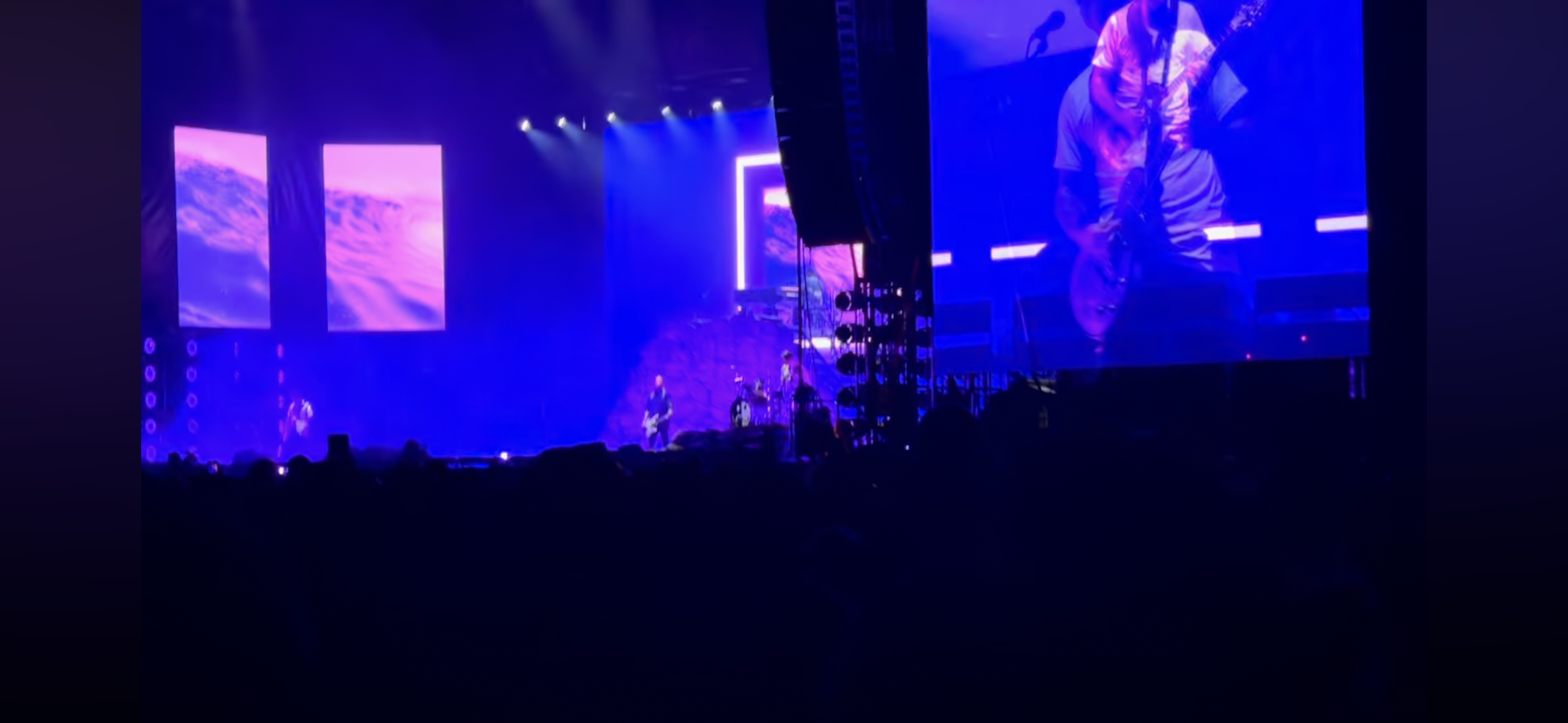 a concert scene with a band performing on stage. the stage is lit with colorful lights, and a screen displays a purple and blue backdrop. the audience is mostly obscured by the darkness, but their silhouettes are visible. the image captures the energy of a live concert and the excitement of the performance.