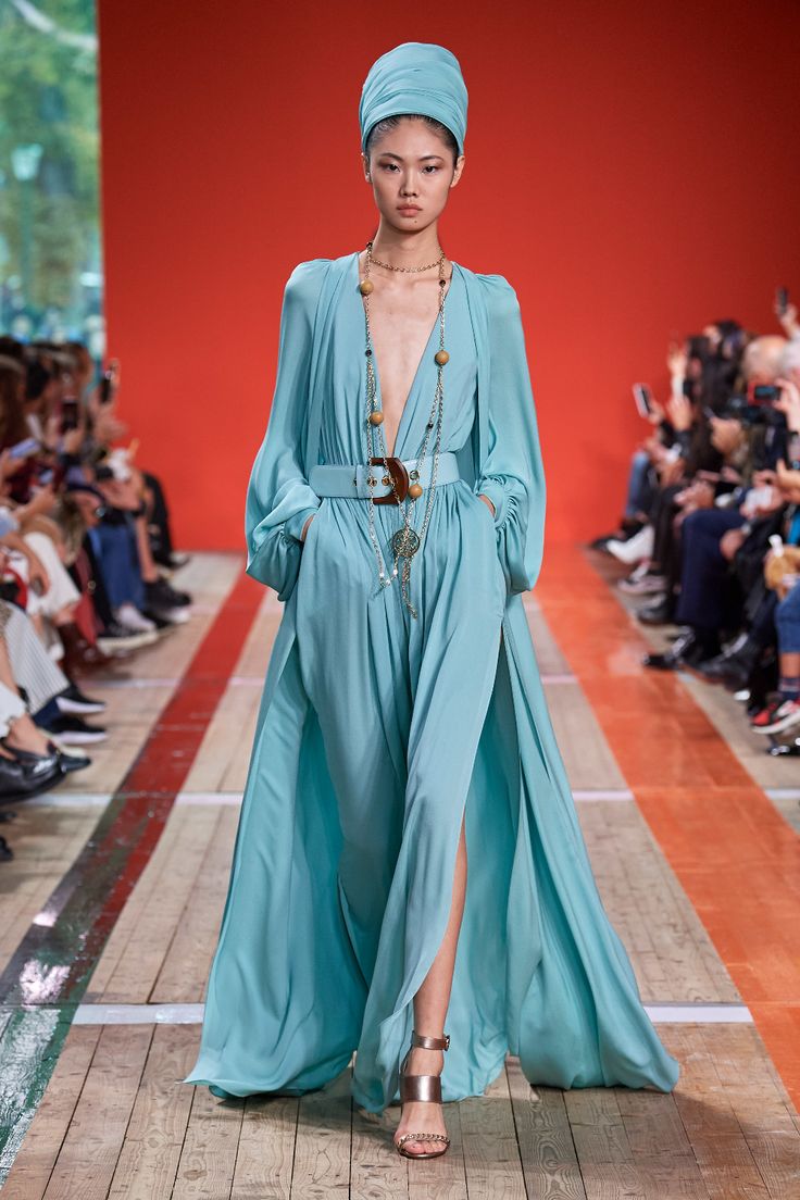 a female model in a long, flowing blue dress with a belt and sandals is walking down a runway. she is accessorized with jewelry including a necklace and a turban hat. there are spectators in the background seated on the floor. the image is of a high fashion show with a glamourous feel.