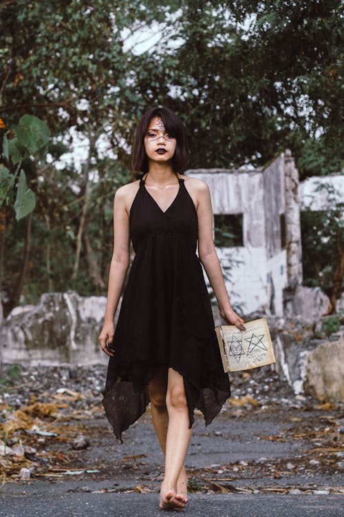 a young woman with dark makeup and a black halter dress walks barefoot on a road surrounded by ruins. she holds a book with pentagrams on its cover. the scene has a witchy or magical atmosphere.