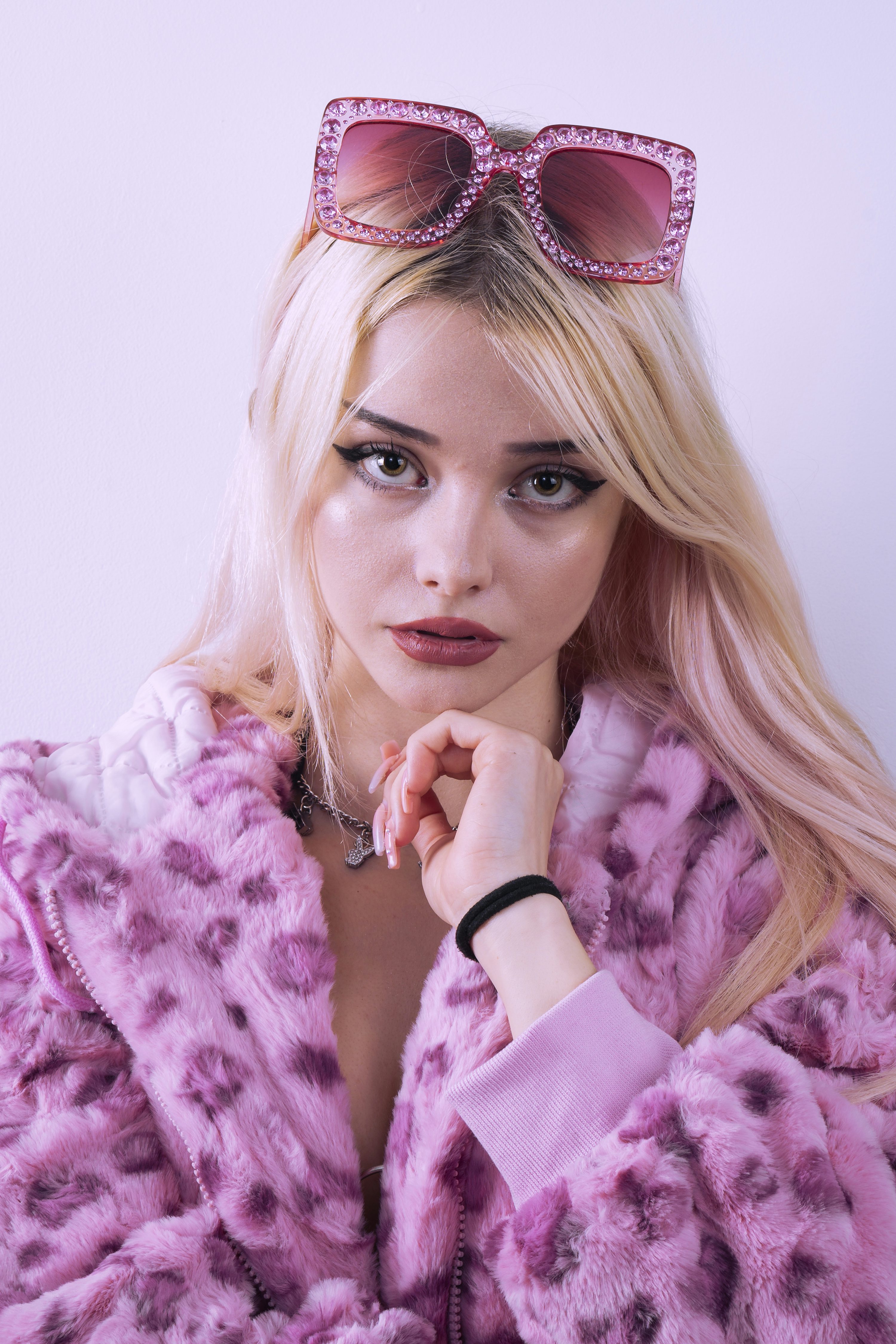 loseup portrait of a young blonde woman wearing a pink leopard print fur coat. she is wearing oversized pink rhinestone sunglasses perched on top of her head, and has subtle makeup.  she has her hand gently resting on her chin. the background is a plain light purple.