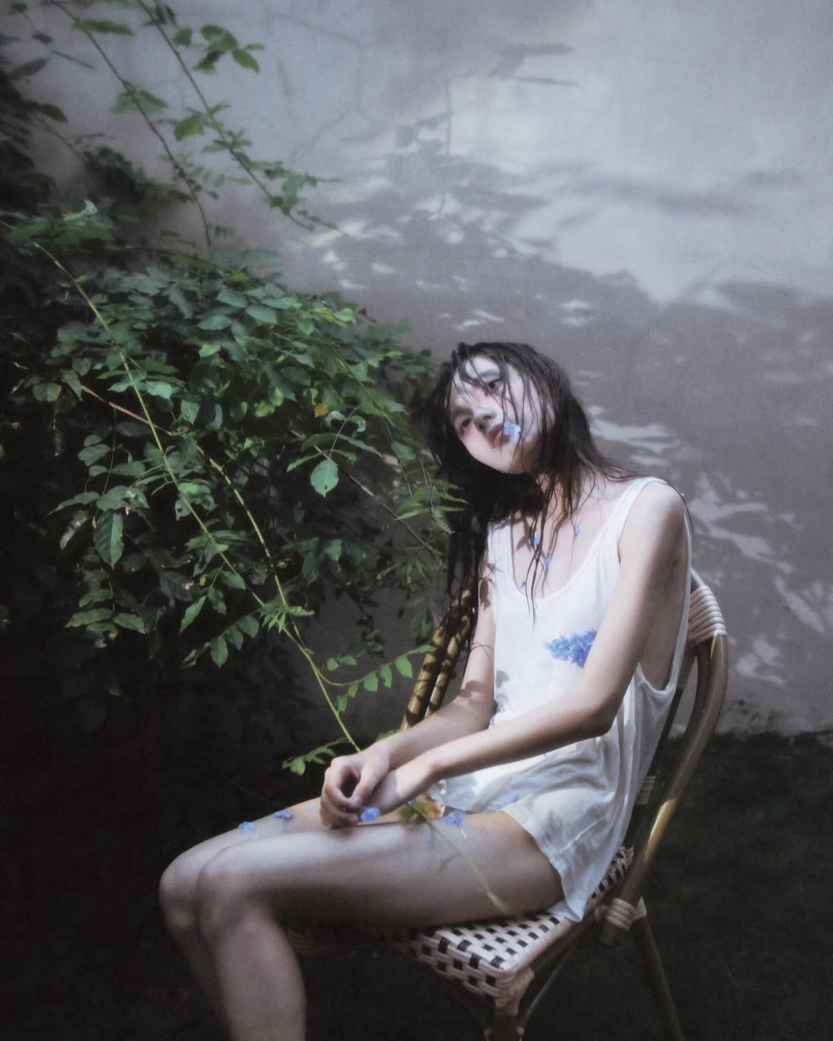 a young woman with dark, wet hair sits on a woven chair in an outdoor setting. she is wearing a white tank top and appears to be wearing a short dress or shorts. small blue flowers are scattered on her body and clothes, as well as placed in her mouth. behind her is a wall with shadows cast by foliage. lush green plants surround her, creating a gardenlike atmosphere. the overall mood is somewhat melancholic and artistic.