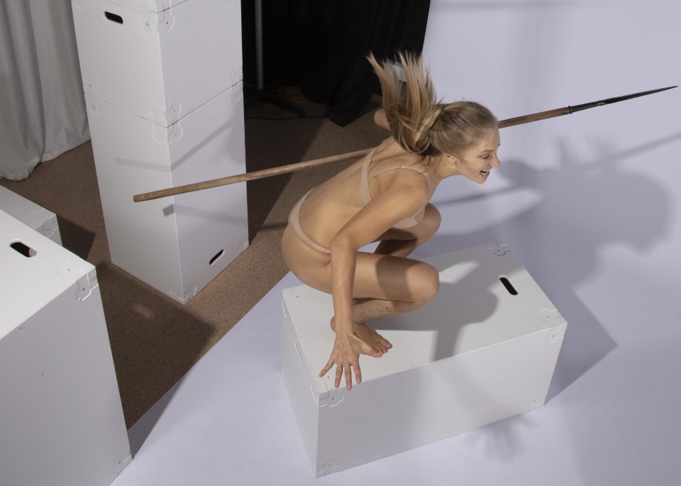 a woman in activewear is jumping over a white box, with a wooden spear positioned across two other boxes, creating a limbo setup in a studio.
