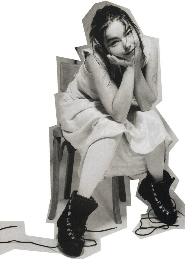 a black and white portrait of a woman sitting on a chair with her arms crossed, wearing a white dress and black boots. she is smiling and looking at the camera.