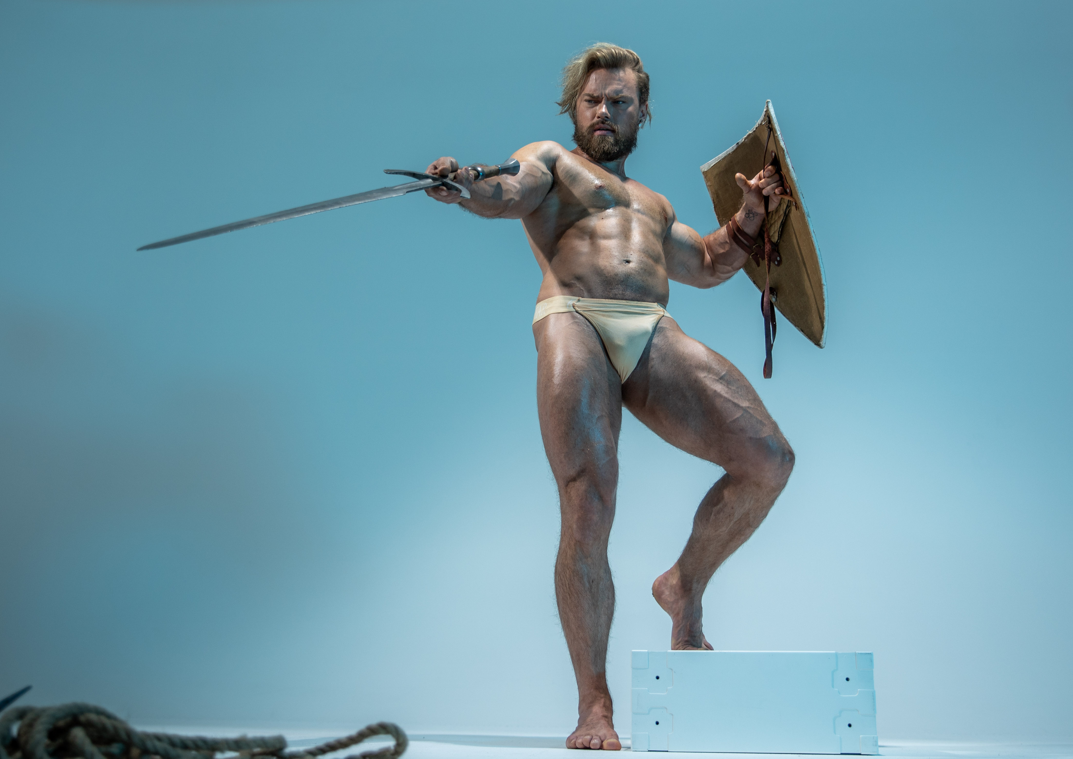 a muscular man with a beard, wearing only a loincloth, stands in a fighting stance on a white platform. he holds a sword in his right hand and a round shield in his left, both seemingly made of metal and leather. he is illuminated by bright, artificial lighting against a light blue background, creating a heroic and powerful image.