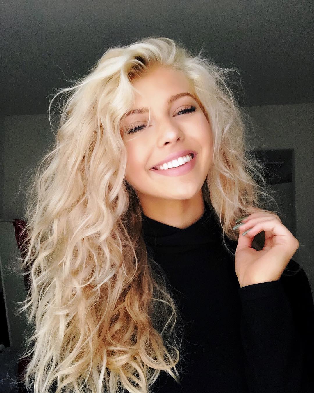 a smiling woman with long, blonde, wavy hair is wearing a black turtleneck shirt and holding her hand up near her face.