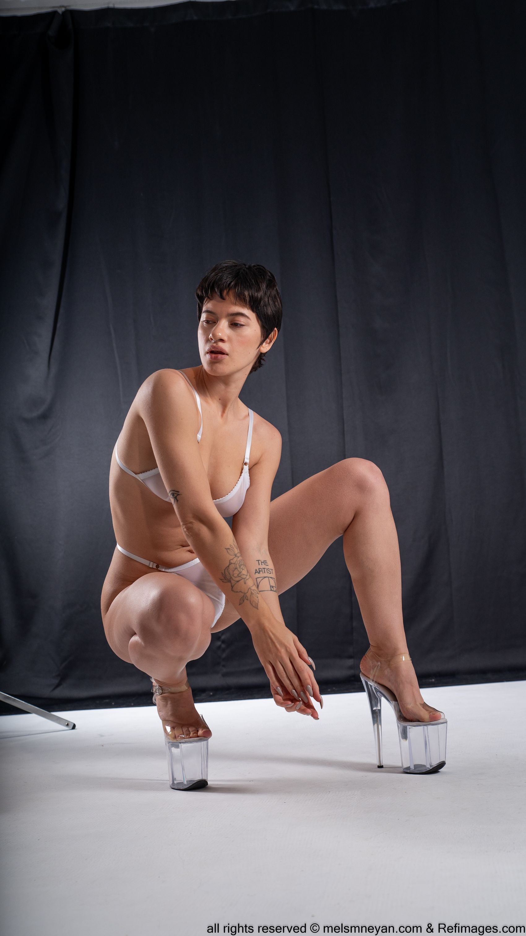 a woman with short, dark hair is squatting indoors in a studio setting. she is wearing white lingerie, including a bra and underwear. she also has on clear platform heels. the woman has multiple tattoos on her arms. she is posing in front of a black backdrop on a white floor. the overall lighting highlights the contours of her body and emphasizes the studio environment.