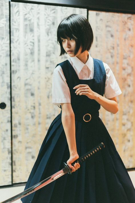 a young japanese school girl wearing a traditional school uniform is standing indoors holding a katana. she is looking away from the camera and her expression is serious. the background is a wooden wall with a pattern.