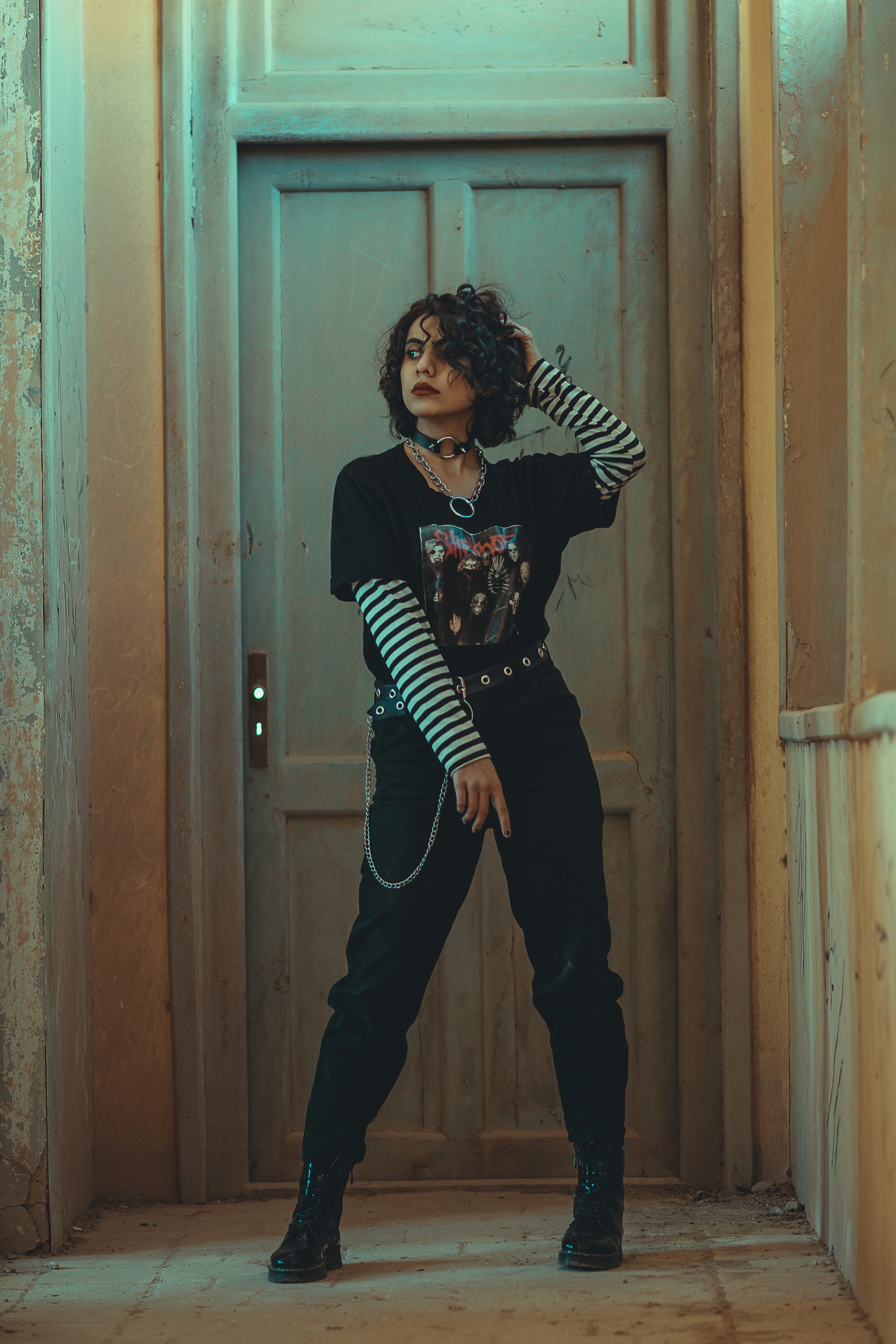 a young woman with curly dark hair is standing in a hallway in front of an old wooden door. she is wearing a black tshirt with a band graphic, black pants, black boots, and a striped longsleeved shirt underneath.  she is accessorized with a choker necklace and a chain. the overall style is grunge or alternative. the setting is slightly dilapidated or aged, adding to the mood of the image.