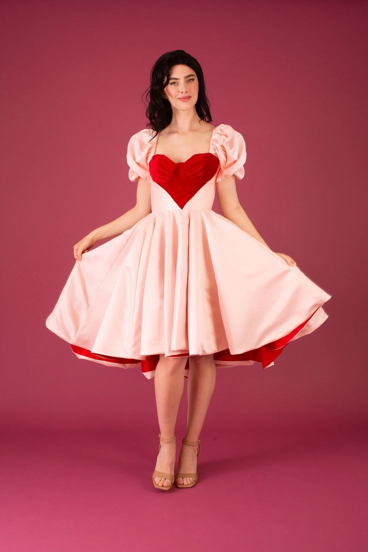 a woman is posing in a pink dress with a red heart on the bodice. she has long dark hair and is wearing strappy heels. the background is a solid pink color. the dress is full and flares out at the waist. the sleeves are short and puffed. the overall look is romantic and feminine.