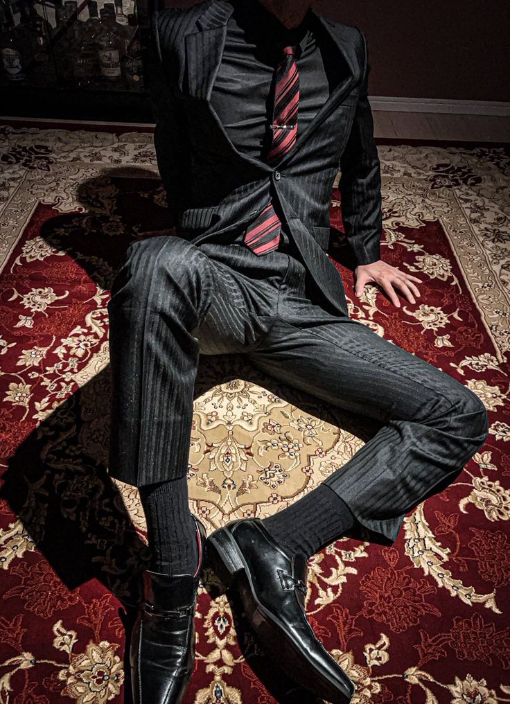 a man in a black suit with a red and black striped tie sits with his legs crossed on a patterned carpet. the carpet has red and beige hues, and the man is wearing black socks and shoes. he is leaning forward with his hands on the floor. the image captures a sense of style, sophistication, and confidence.