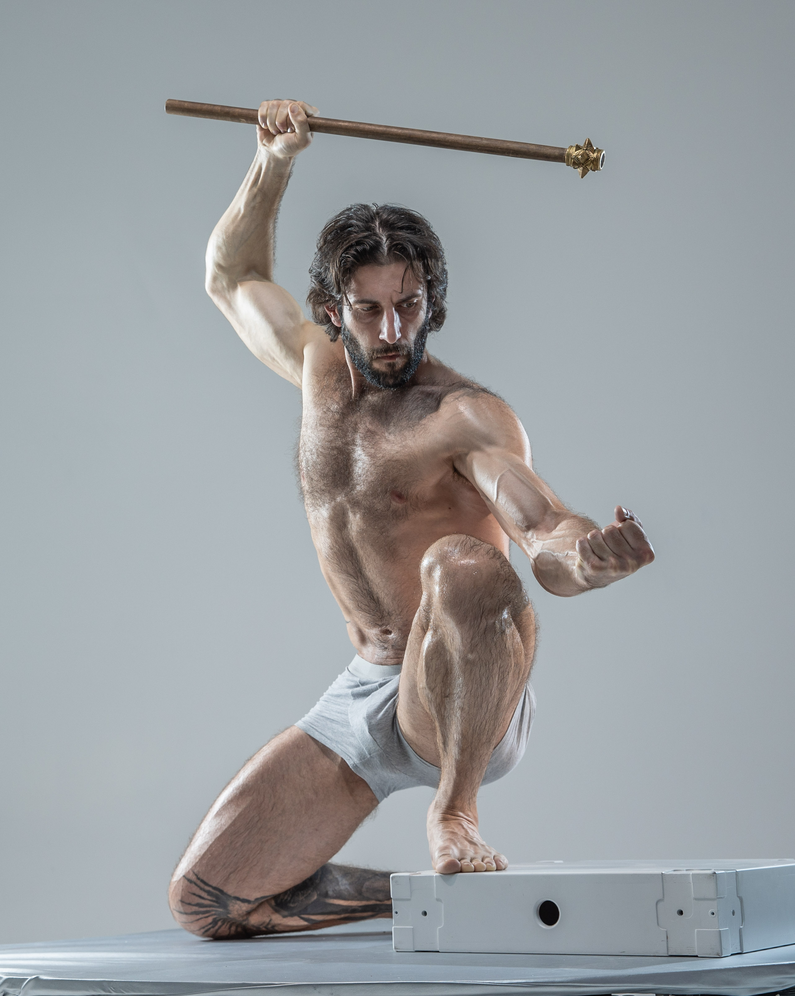 a muscular, shirtless man with long dark hair and beard assumes a dynamic fighting stance against a neutral gray background. he grips a wooden staff with a metal tip, his body tensed in anticipation of attack or defense. the image emphasizes his physique and the intensity of the moment, captured with studio lighting that highlights his muscles and the motion blur of his arm. he kneels on one leg upon a white rectangular platform, which contrasts with the plain backdrop.