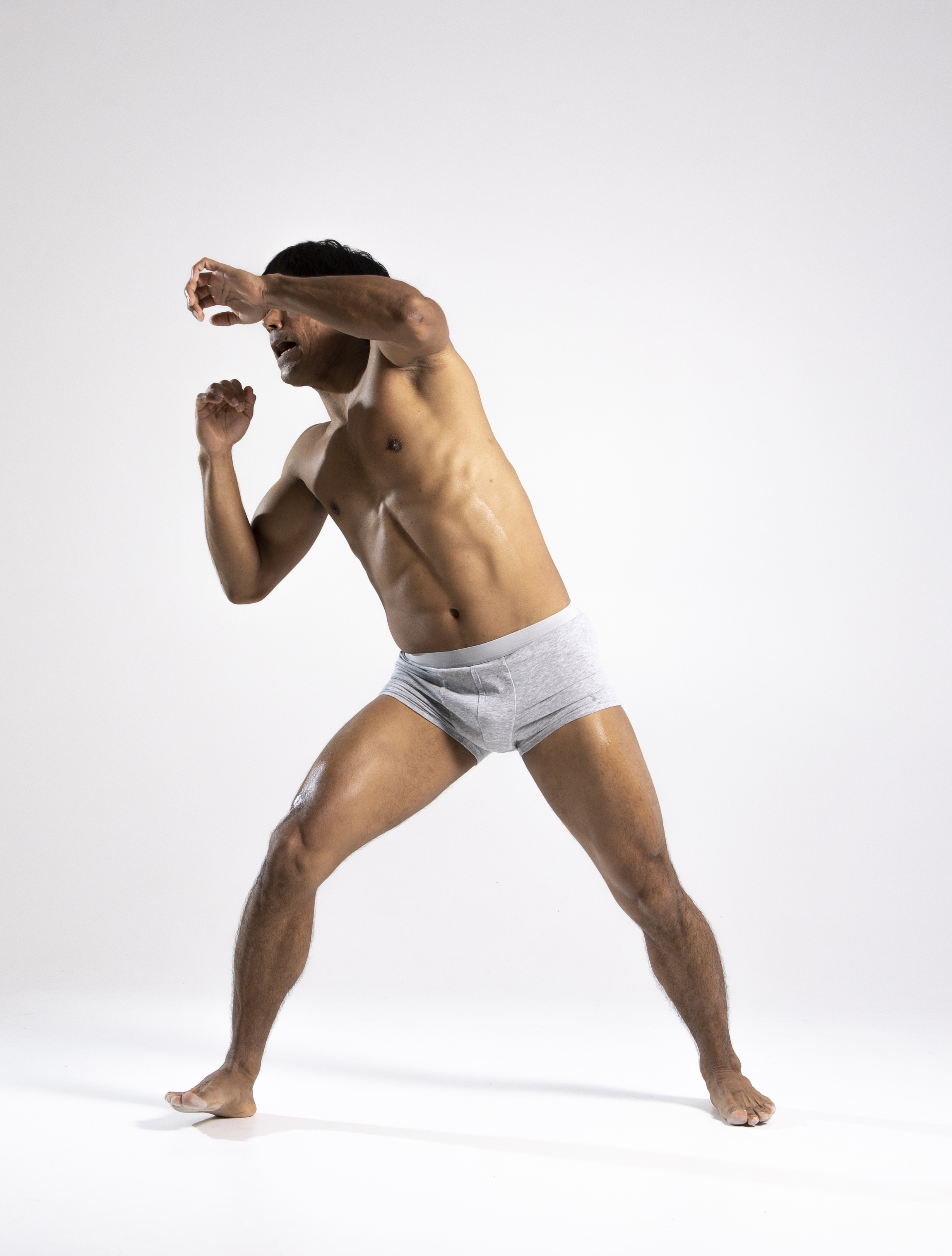a shirtless man in gray underwear is captured in a dynamic fighting stance against a plain white background. he is barefoot with his legs spread apart, knees slightly bent, showcasing a defensive posture. his fists are raised near his face as if guarding against an attack. the man's muscular physique is emphasized by the lighting, highlighting the tone of his skin and the contours of his body. he exudes a sense of agility, determination, and health. the studio setting directs focus entirely on the subject and his athletic form.
