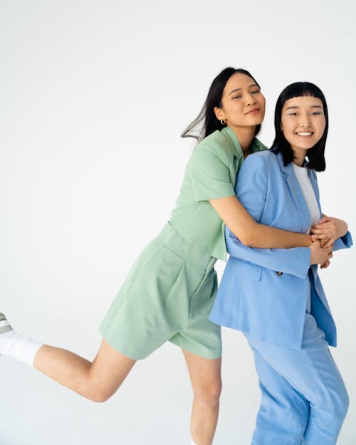 the image shows two young asian women joyfully embracing each other against a white background. one woman is wearing light green shorts, and the other is wearing a light blue suit.  they appear happy and relaxed, conveying a sense of friendship and closeness. the overall mood is light and cheerful, emphasizing the connection between the two individuals.  the pose is dynamic, with one woman playfully leaning on the other.