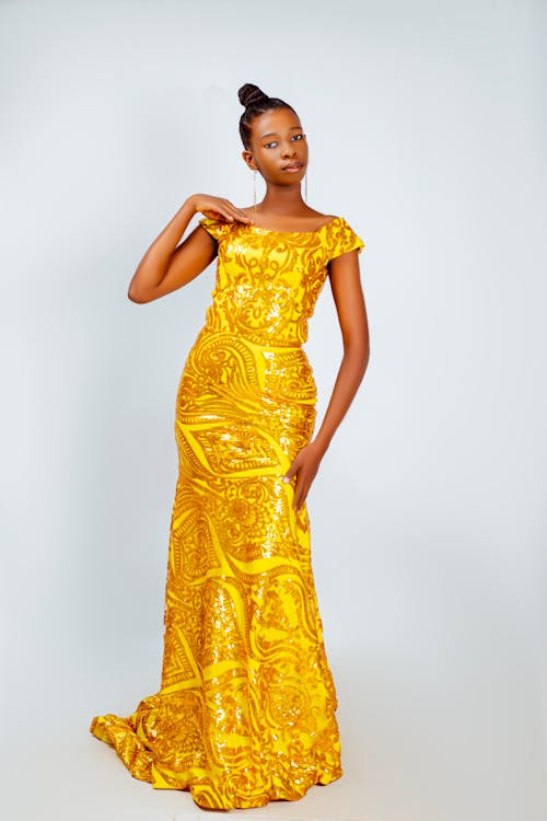 a slim, elegant african woman poses gracefully in a studio shot. she is wearing a shimmering, bright yellow evening gown embellished with sequins and intricate patterns. her hair is neatly styled in a high bun, and she accessorizes with simple, long earrings. she exudes confidence and sophistication as she places a hand delicately on her shoulder.
