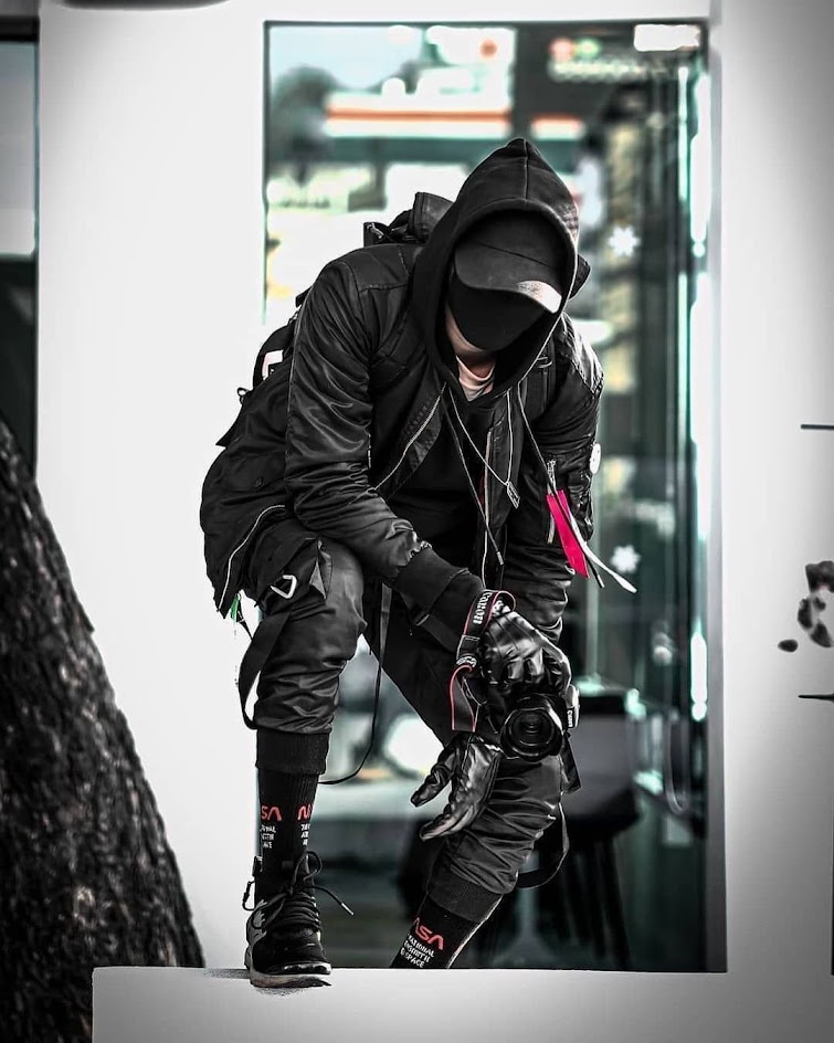 a man wearing all black clothing is holding a camera. he has a black mask on and is standing in front of a glass door. the man is dressed in a street wear style with an edgy fashion sense. he has sunglasses on, and he looks cool and stylish. he is a photographer who is ready to take some amazing photos.