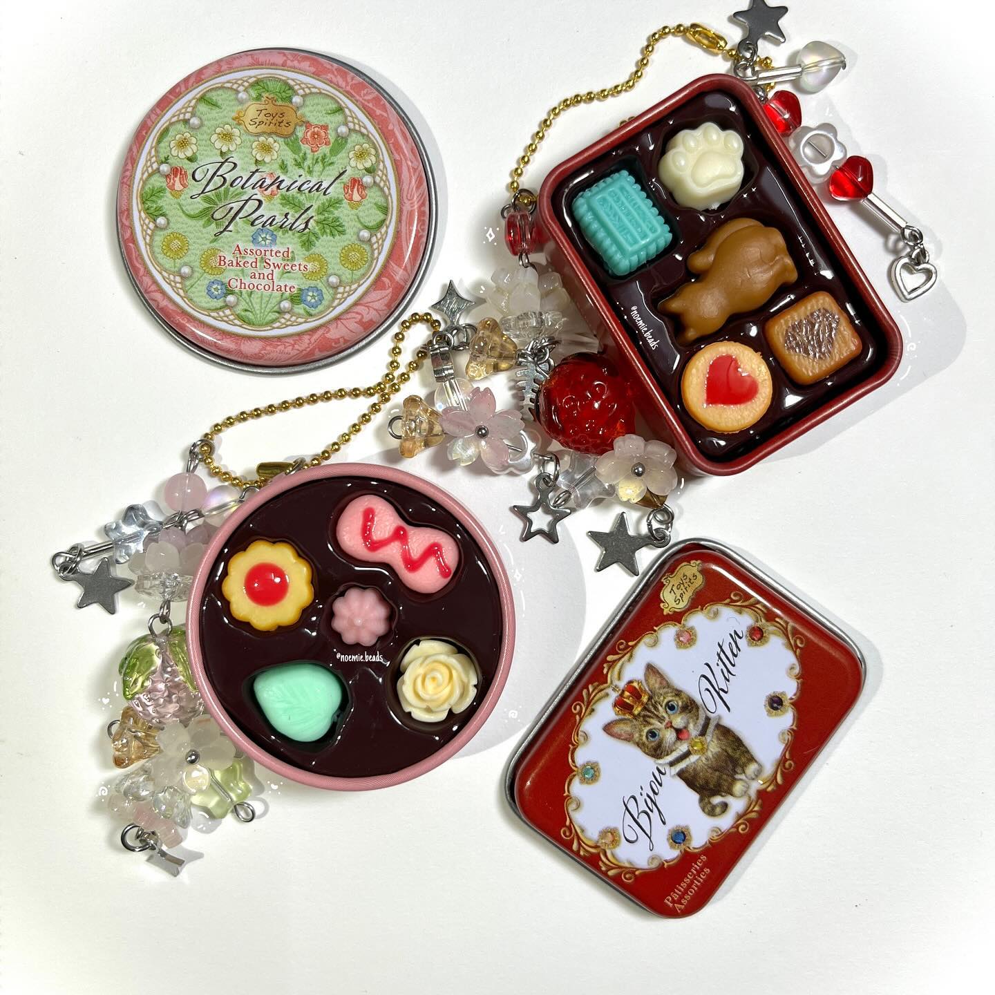a collection of miniature dessertthemed trinkets are displayed on a white surface. the items include a round tin labeled "botanical pearls assorted baked sweets and chocolate", a rectangular tin filled with miniature chocolates, a round trinket resembling a chocolate assortment with several small desserts, and a rectangular tin featuring a cat. all the trinkets have beads and charms attached, including flowers, stars, and other decorative elements.