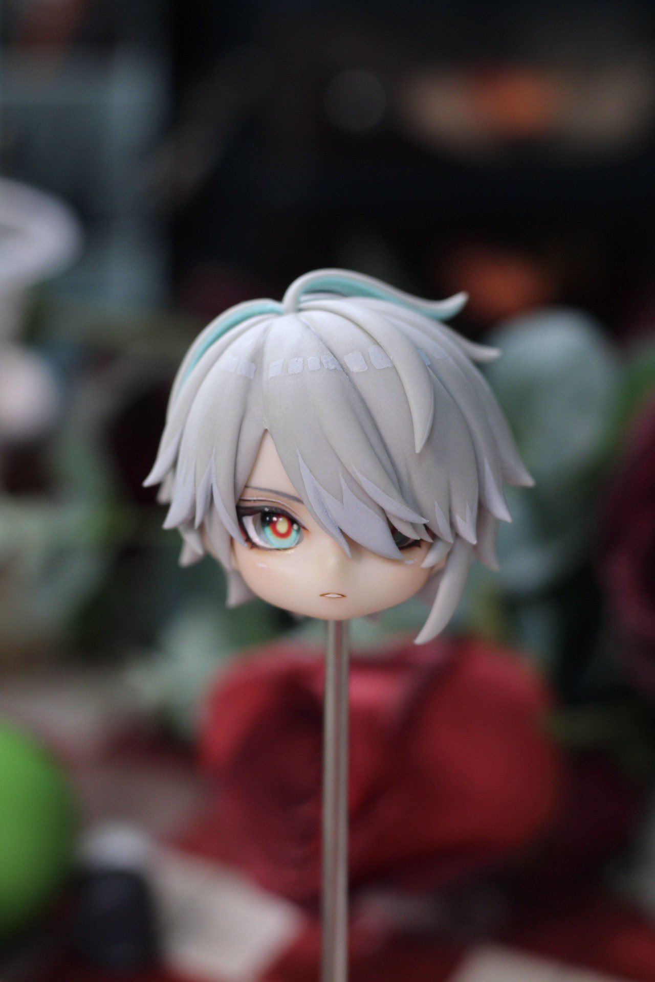 a close up shot of a handmade anime figurine head with white hair and red eyes. the head is placed on a wooden stick. the background is blurred and shows red fabric and green foliage. the figurine is cute and wellmade.
