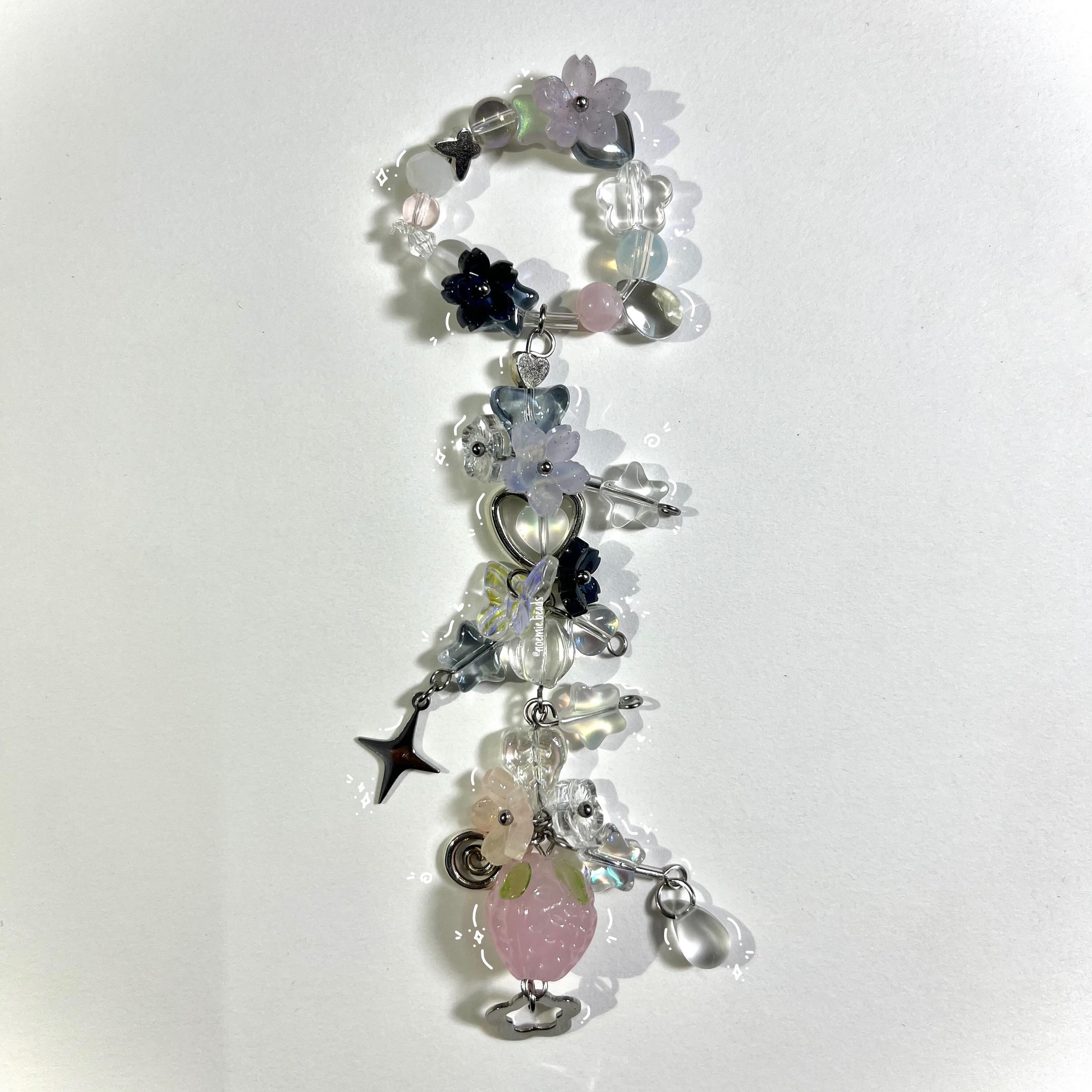 a beaded keychain or pendant featuring a variety of charms and beads in pastel colors. the piece includes floral beads, clear and colored beads, and metal charms such as a star and a heart. the arrangement is vertical, with the beads and charms dangling from a beaded bracelet at the top.