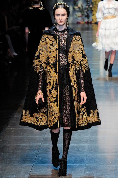 a runway model walks down the catwalk wearing a striking black cape adorned with intricate gold embroidery.  the cape features a floral and baroqueinspired pattern and is worn over a black lace dress. the model is accessorized with dark socks and stylish ankle boots. her dark hair is neatly pulled back, and her makeup is minimal and elegant. the background shows other models and blurred spectators indicating a fashion show setting. the overall ambiance is one of high fashion and luxury.