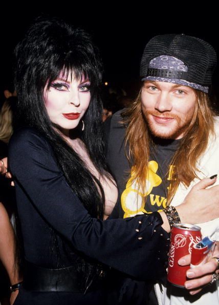 this image features a woman with black gothicstyle dress and a man in a white tshirt and a black baseball cap. the woman has long black hair, while the man has long blonde hair.  the man is holding a can of soda and they both appear to be posing for a picture.  they are both looking at the camera.  the background is blurry, but it appears to be a concert or music festival.