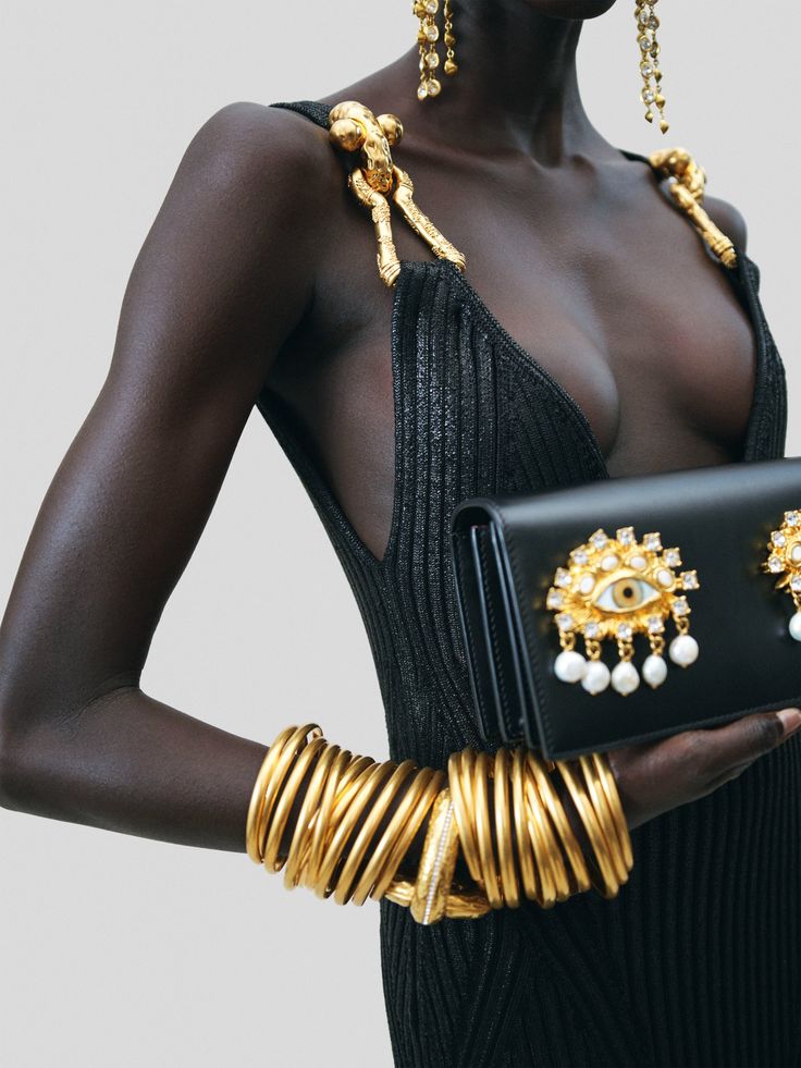 a woman wearing a black dress with gold jewelry, including bracelets and earrings. she is holding a black clutch with a gold eye design.