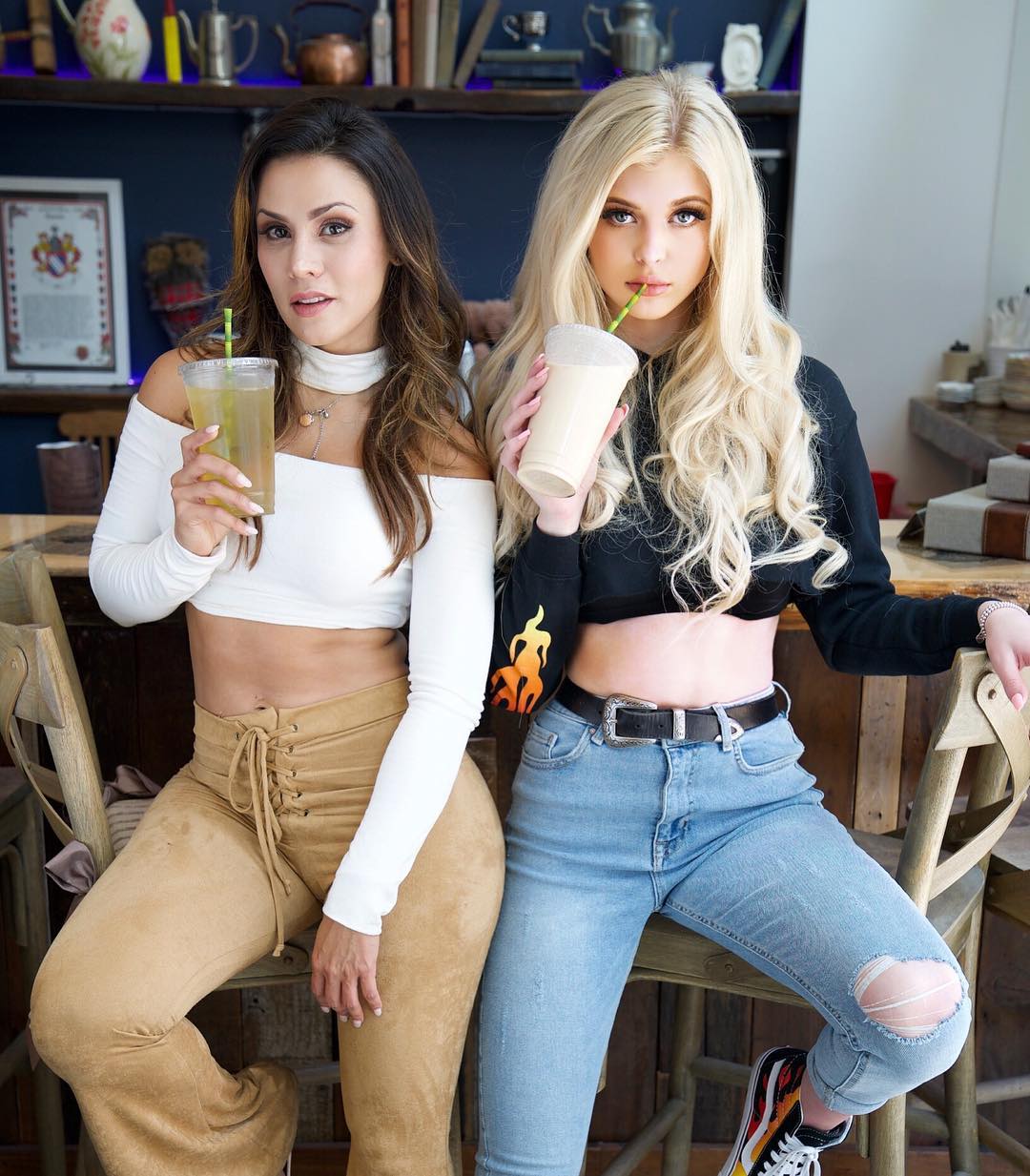 two women are sitting on stools in what appears to be a lounge bar. the woman on the left has brunette hair and is wearing a white offtheshoulder crop top and tan highwaisted fronttie pants. she's holding a clear plastic cup with a yellow drink. the woman on the right has blonde hair and is wearing a black crop top with flame designs on the sleeves, highwaisted ripped jeans, and a black belt. she's holding a white drink in a clear plastic cup. both women are looking at the camera.