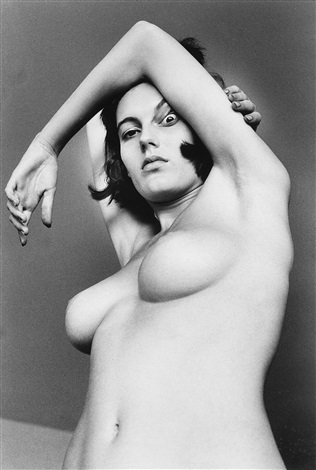the photograph is a black and white, closeup portrait of a nude woman. her body is turned slightly away from the camera, with one arm gracefully draped over her head and the other resting on her hip. the lighting emphasizes the curves and texture of her skin.  her expression is serious and somewhat aloof. the overall style is reminiscent of classic art photography, possibly from the mid20th century, with a focus on form and composition. a dark element seems to be partially obscured near her temple.
