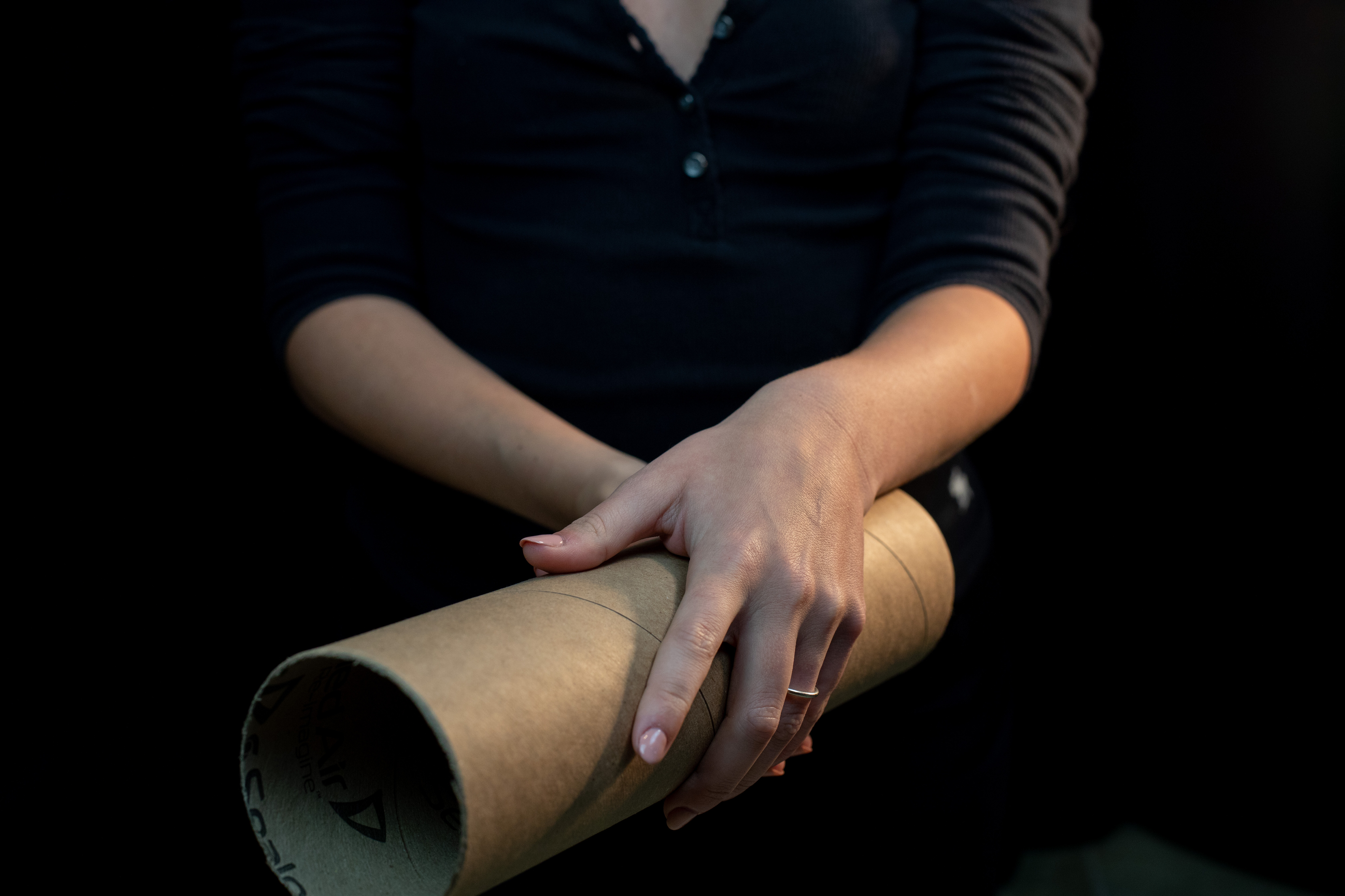 a closeup image of a person's hands holding a cardboard tube. the tube is made of brown kraft paper and has a seam running down the side. the person's hands are clasped around the tube, and their nails are painted a light color. the background of the image is dark.
