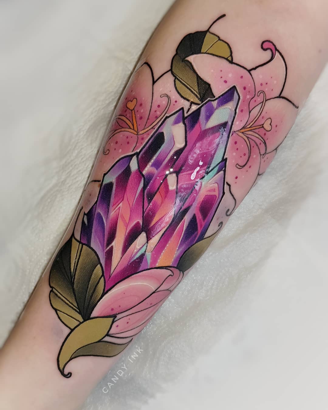 a neotraditional style tattoo on a person's forearm. the tattoo features a large, multifaceted crystal in shades of purple and pink, surrounded by pink lily flowers with green leaves.