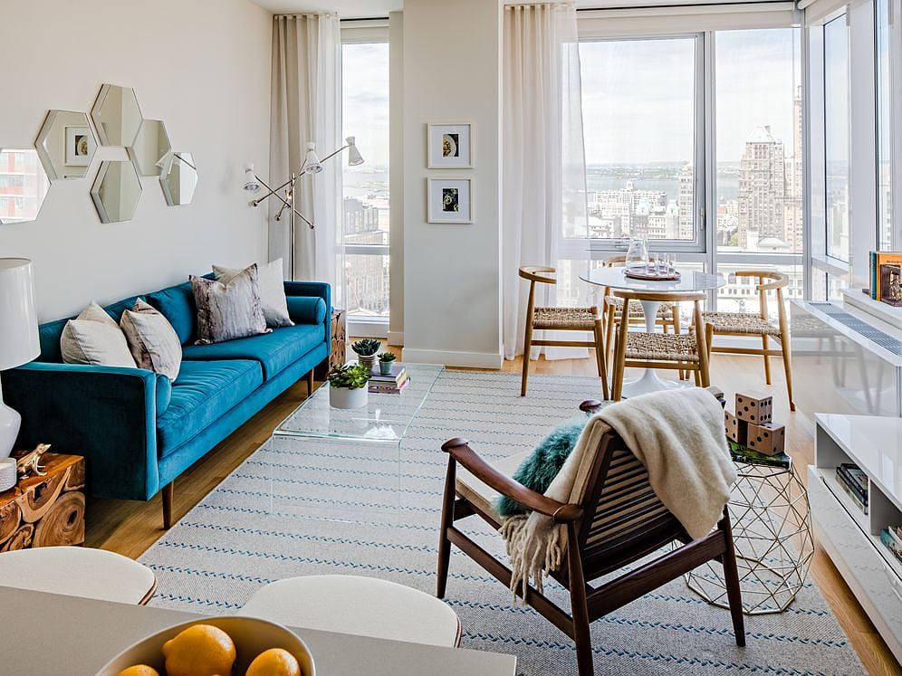 a modern living room with a teal blue sofa and wooden chairs. the living room has large windows with a view of the city.  the floor is covered in a patterned rug and there is a glass coffee table in the center of the room. the room is decorated with minimalist decor.