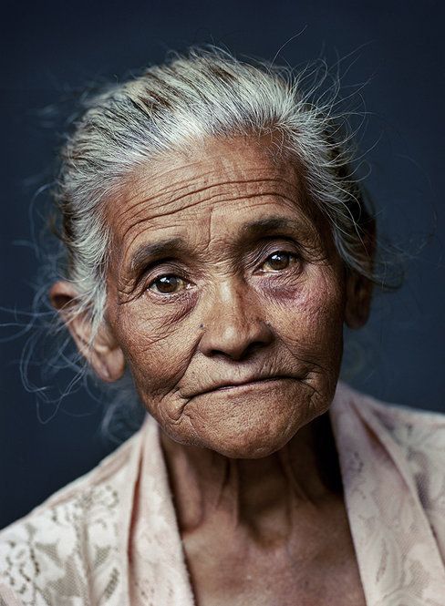 a closeup portrait of an elderly woman with gray hair and deeply lined skin. she has brown eyes and a slightly sad or thoughtful expression. she is wearing a light pink patterned garment. the photo highlights the details of her weathered face, showcasing the wrinkles and textures of aging.