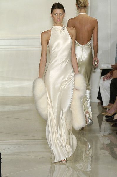 a fashion show features a model walking the runway. she wears a white, sleeveless gown and white fur sleeves. another model wearing a similar gown walks behind her.