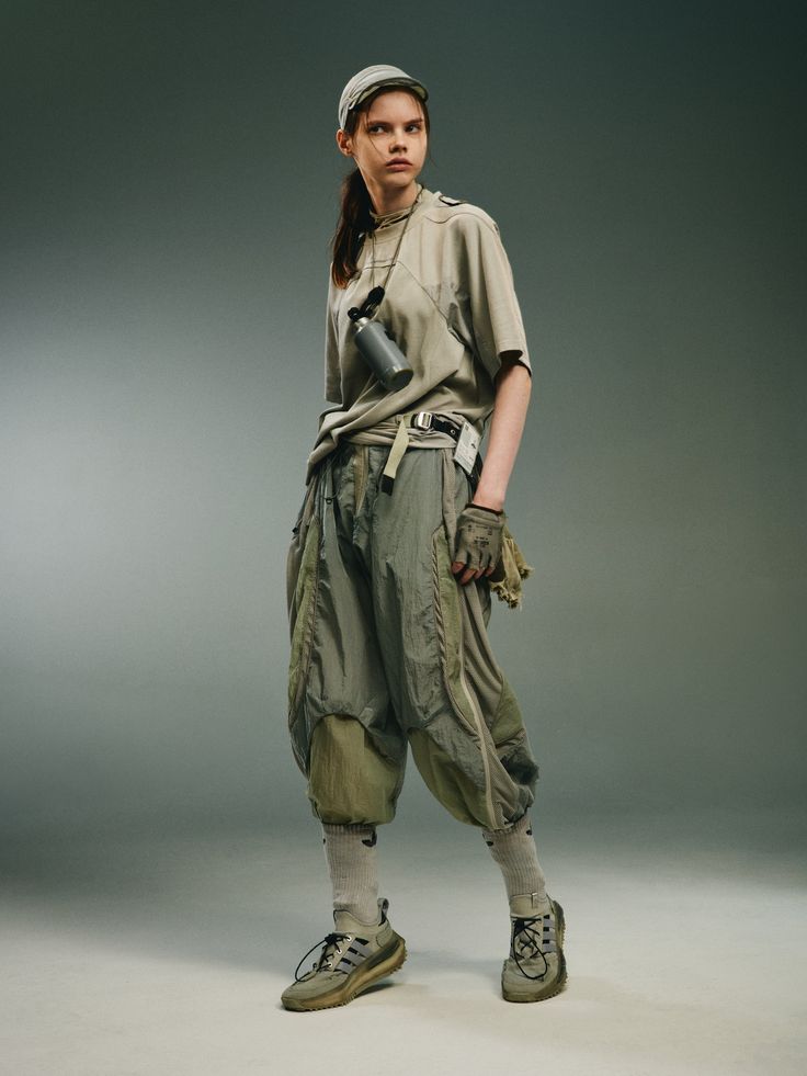 the image showcases a female model in a stylish, contemporary outfit. the model is wearing a light beige oversized tshirt, loose olivegreen cargostyle pants that are cropped above the ankle, light beige sneakers, a beige baseball cap, and short socks. several accessories include a small grey water bottle,  a fabric belt, and gloves, adding to the overall utilitarian yet fashionable look.  the model is posing in a relaxed yet controlled manner against a plain grey studio background. the overall aesthetic is minimalist and sophisticated. the photo appears to be taken for a fashion editorial or a highfashion lookbook.