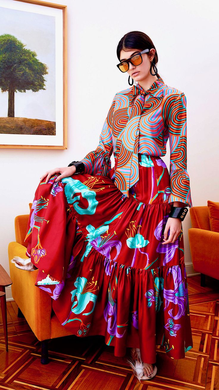 a full shot of a fashion model sitting on a chair in a room with a hardwood floor. she is wearing a striking outfit: a red floral print tiered midi skirt paired with a retrostyle multicolored blouse with swirling patterns. the blouse has long sleeves and a tied neck.  the model wears sunglasses, earrings, and what appears to be a bracelet or cuff on her wrist. the overall style is elegant and vibrant. the background includes an artwork on the wall and orange armchairs. the image has a welllit and stylish feel, suggesting a fashion editorial or campaign.