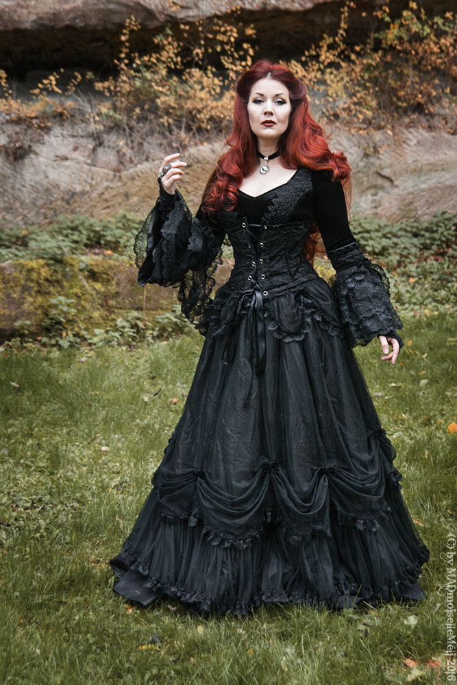a woman with red hair stands in a grassy outdoor setting, wearing a floorlength black gothic dress with lace detailing, long sleeves, and a corset. she wears a choker and a ring. the background includes trees and natural landscape.