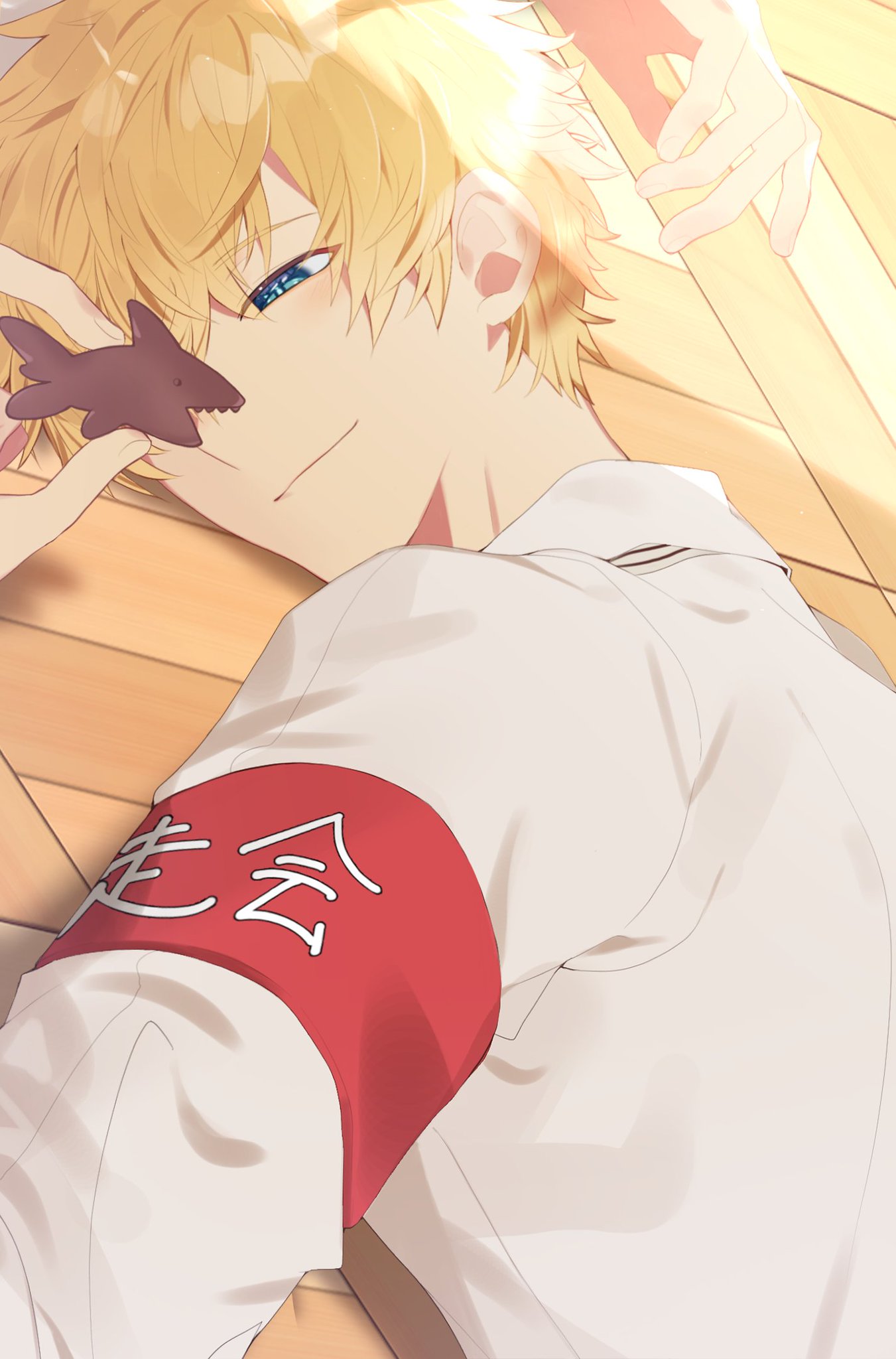 a cute anime boy with blond hair and blue eyes is lying on a wood floor. he is smiling and holding a cookie in his hand.  he is wearing a white shirt with a red band on his arm. the band has chinese characters on it. there is a faint outline of a hand in the background. the boy is holding a shark cookie.