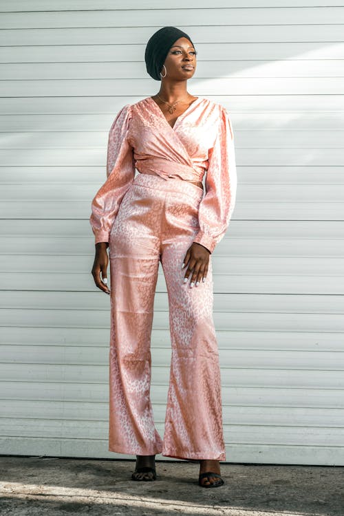 a fullbody shot of a stylish woman posing outdoors against a white wall. she is wearing a pale pink satin jumpsuit featuring a leopard print. the jumpsuit consists of a wrapstyle top and wideleg pants.  the woman also sports a black headwrap or turban. she is standing with a confident pose, her gaze directed slightly away from the camera. the overall style is chic and elegant.