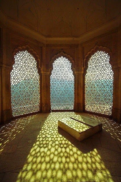 the image shows the interior of a stonebuilt room in amber fort, jaipur, india. the room features three arched windows with intricate latticework that casts a beautiful pattern of sunlight and shadows on the floor. a raised rectangular stone platform sits in the center of the room, also bathed in the patterned light. the warm, earthy tones of the stone and the interplay of light and shadow create a serene and captivating atmosphere.