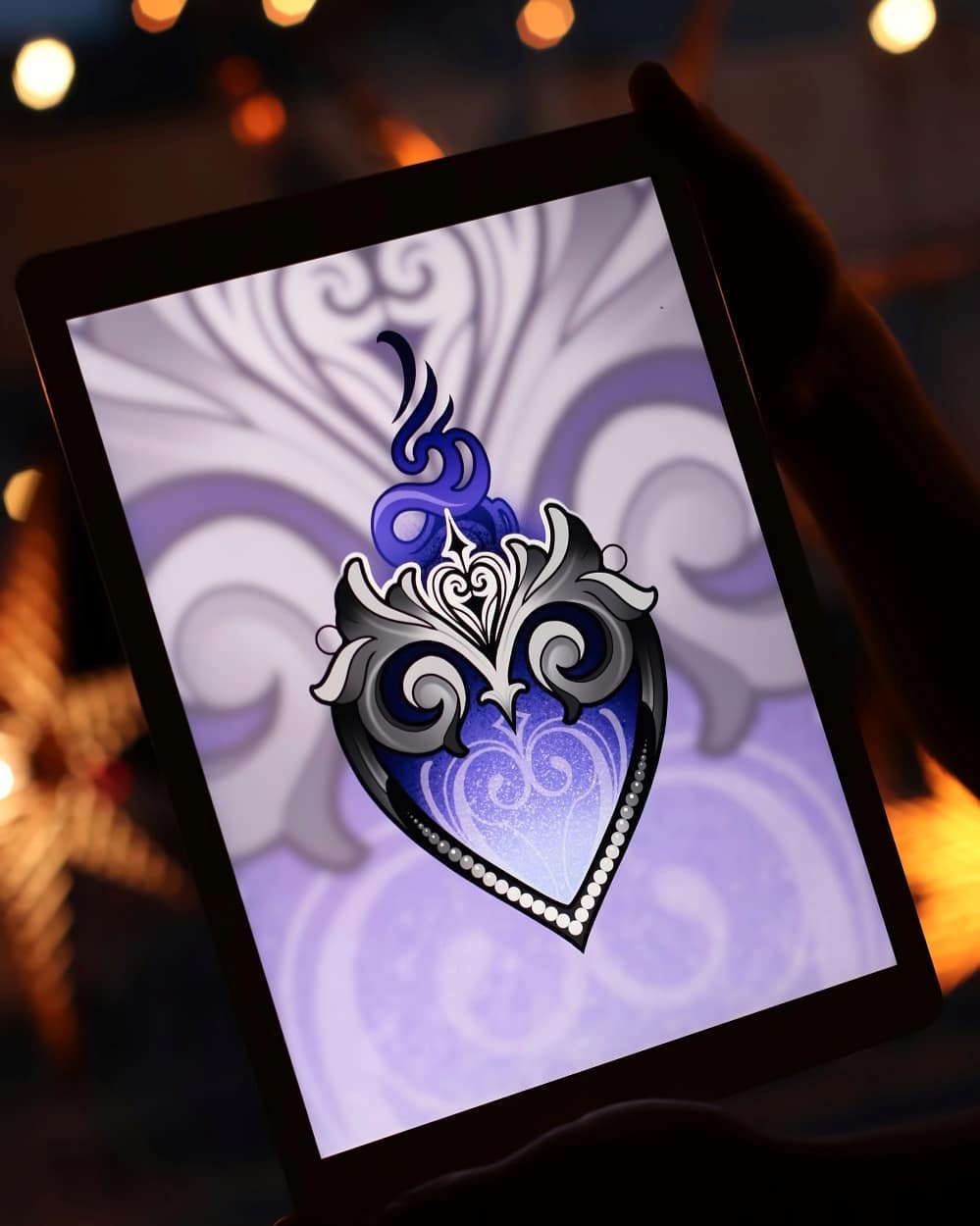 a tablet is being held by a hand. the screen displays a digital drawing of an ornate heart with blue and silver detailing on a purple background. the image is brightly lit, possibly by the light from the screen itself. the hand holding the tablet is silhouetted against the bright screen, making it difficult to discern any details.  the overall aesthetic suggests that the image displayed might be a tattoo design.