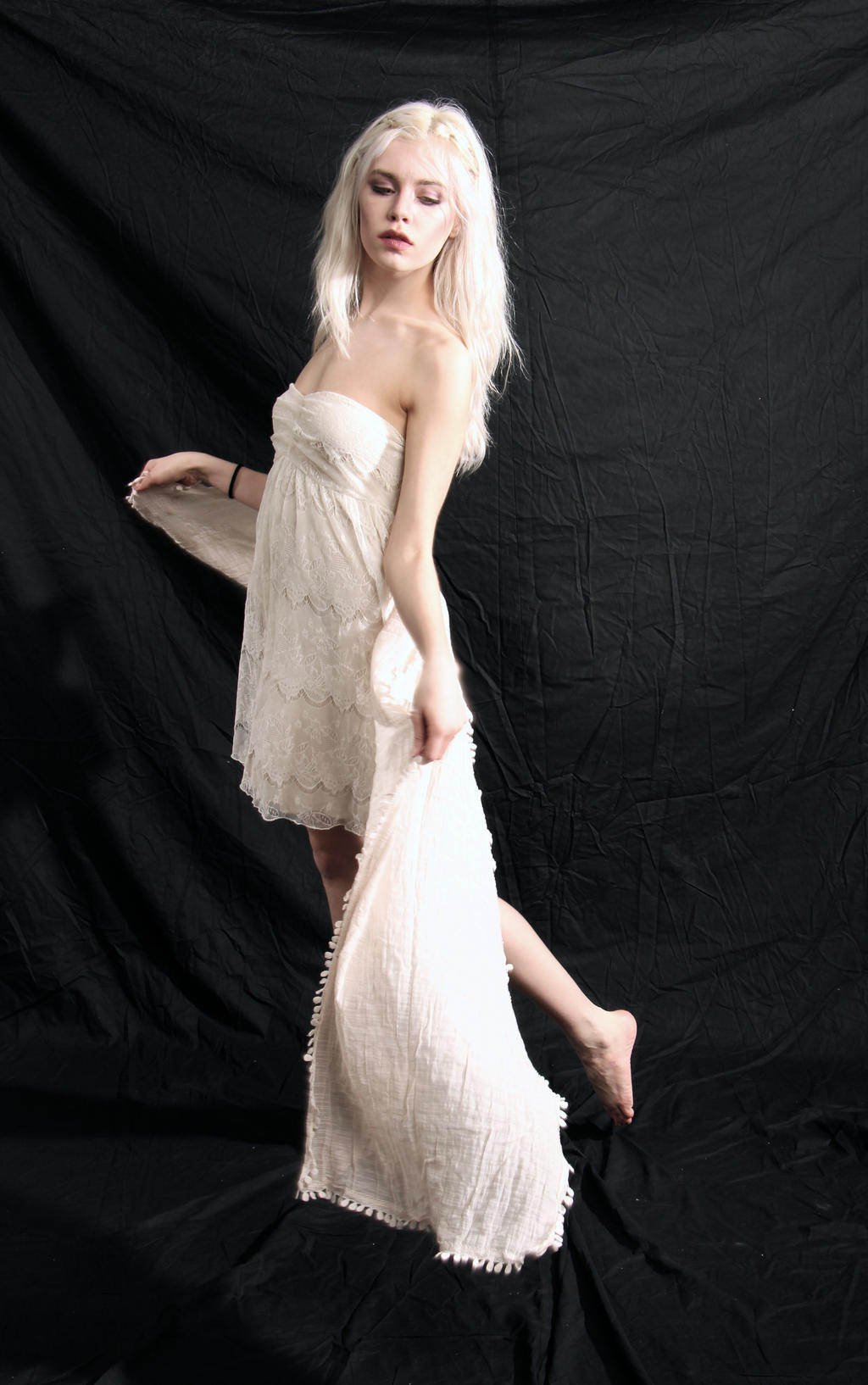 a full shot of a young woman with long, platinum blonde hair, wearing a white lace strapless dress and a sheer white scarf, poses gracefully against a black backdrop. her posture and the flow of the fabric create a sense of ethereal elegance and movement. the contrast between her pale skin and the dark background accentuates her features and the details of her dress.  the image has an artistic and dreamy quality.