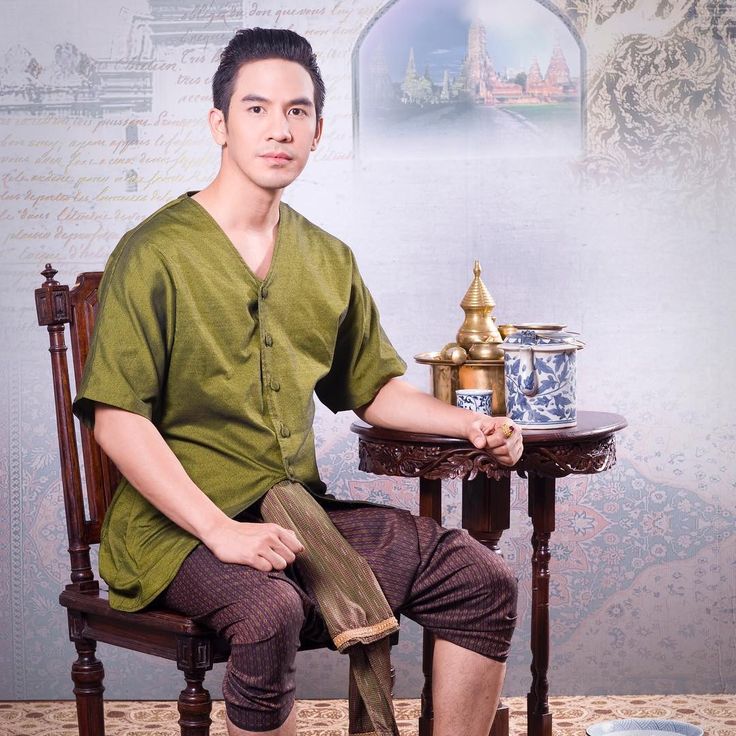 a handsome man in traditional thai clothing is sitting in a chair and smiling. he is wearing a green shirt and brown pants. he is sitting at a table with a teapot and cups. the background is a blurred image of a thai temple.