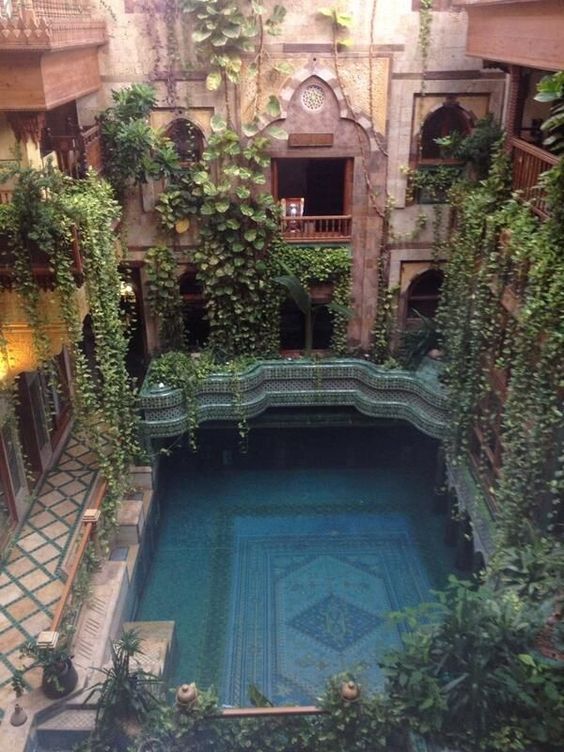 a highangle shot reveals a striking indoor swimming pool within a courtyard setting, heavily adorned with lush greenery. the pool features an intricate mosaic pattern at its base and is surrounded by tilework. the architecture includes archways, windows, and a balcony, with plants and vines cascading down the walls. the design suggests a moroccan or islamic style.
