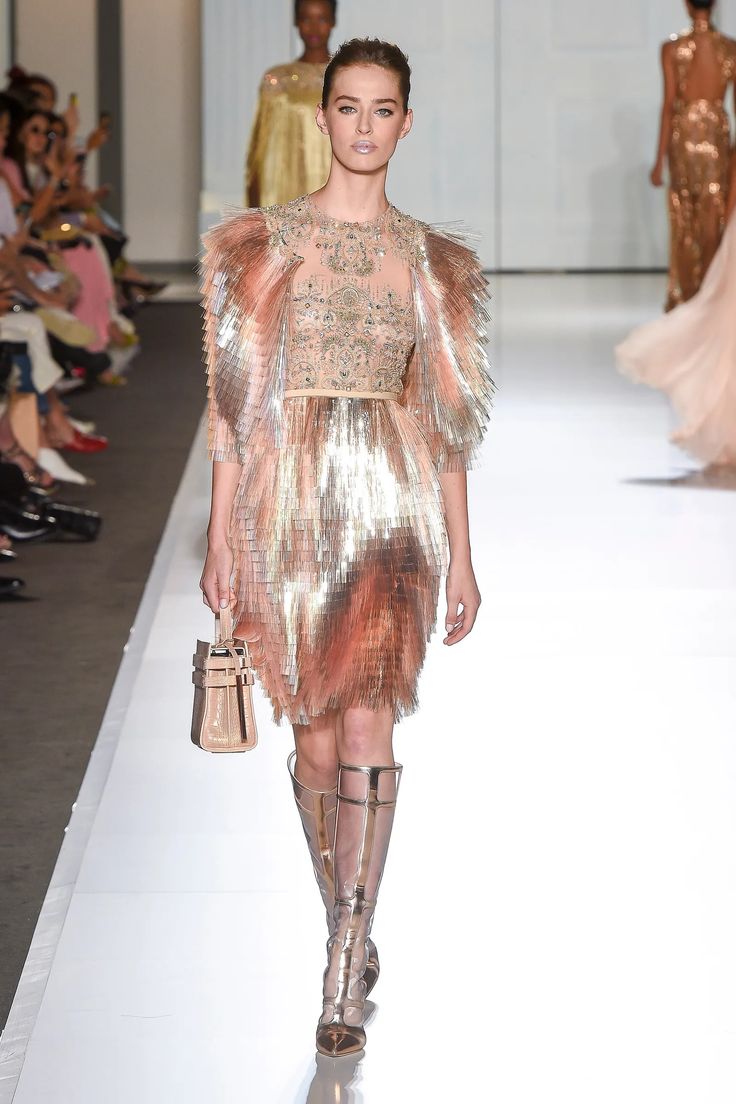 a model walks the runway in a gold and pink sequined dress with large shoulder ruffles. she is wearing highheeled gold boots. she is holding a small gold purse in her hand. she is wearing her hair pulled back and her makeup is done in a natural, glowy style. the background is a white wall, and the model is walking towards the camera.