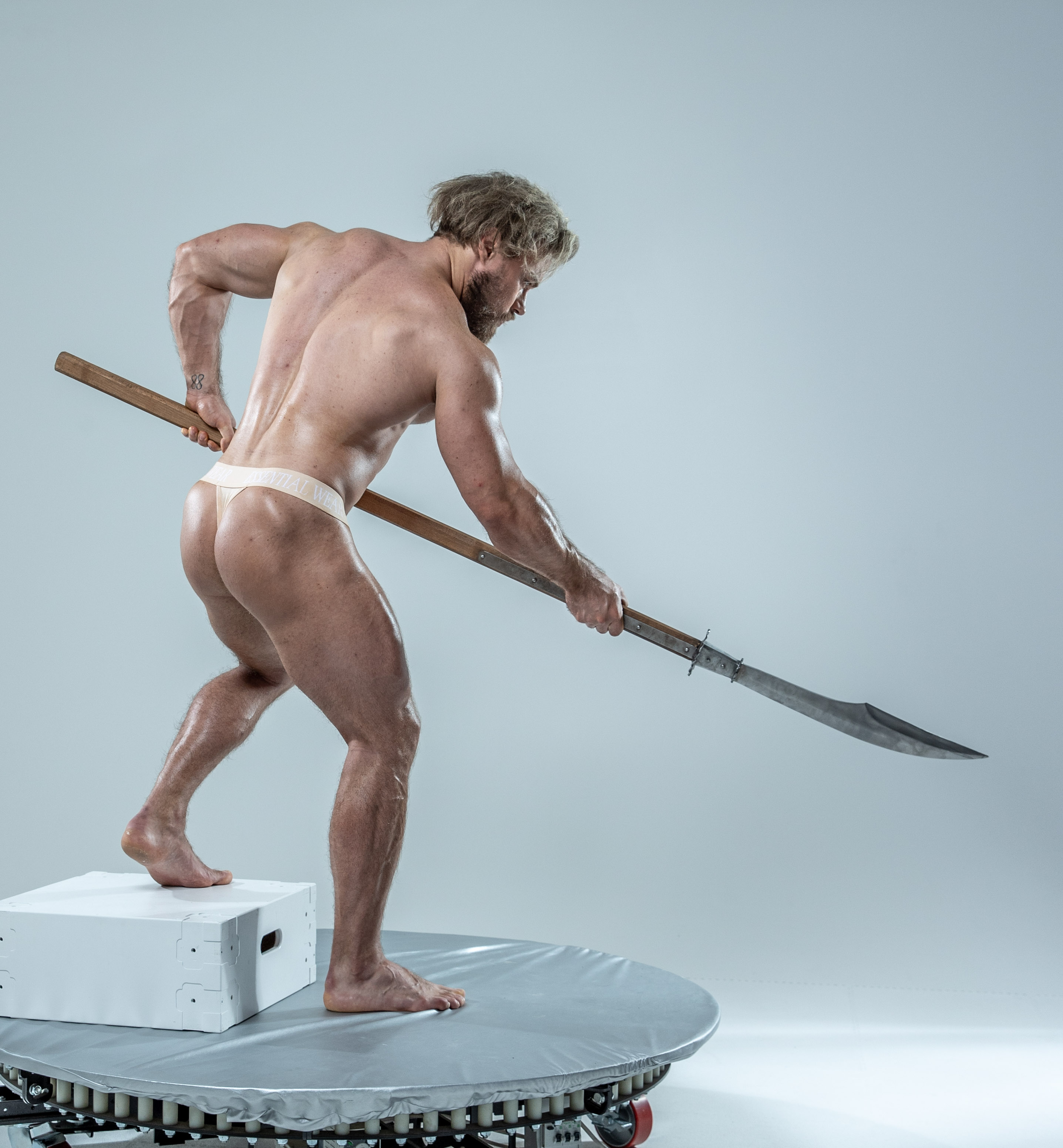 the image showcases a muscular man in a dynamic pose, wielding a spear. he is positioned on a rotating platform within a studio environment, suggesting an artistic photoshoot or study of movement. the soft lighting emphasizes his physique and the spear's form. his stance suggests impending action, capturing a moment of poised strength.